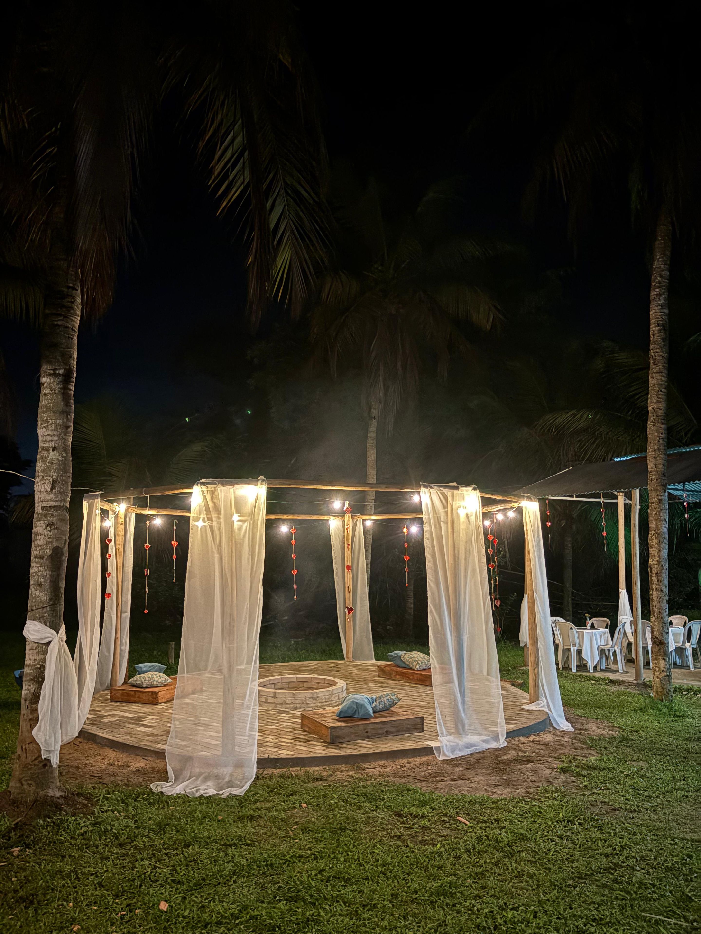 An outdoor seating area with a wooden bench and a small table, surrounded by white curtains and string lights.