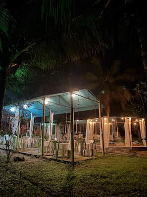 An outdoor dining area with tables and chairs, a covered seating area, and a backdrop of palm trees. - Pousada Recanto Mirante das Águas (União dos Palmares)