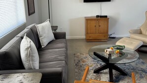 A living room with a sofa, a glass coffee table, a television mounted on the wall, a wooden cabinet, and a beige armchair. - Sweeping Desert & Mountain Views Near Downtown Joshua Tree & JT National Park (Joshua Tree)