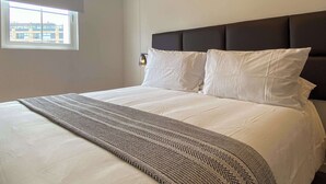 Quarto de hotel com uma cama grande, cabeceira, janela e luminária fixada na parede.