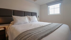 Una cama bien hecha con sábanas blancas y un plaid estampado, un cabecero oscuro, una mesita de noche con lámpara, y una ventana con persianas.