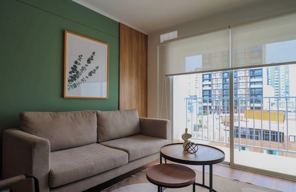 A modern living room with a sofa, a small round table, and a large window offering a view of a cityscape. - w* | Curated Urban 2BR w/ Balcony in Miraflores (Miraflores)