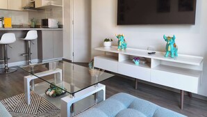 Un soggiorno moderno con un tavolino in vetro, un divano, una TV a parete e un'area cucina sullo sfondo.