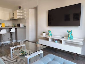 Ein modernes Wohnzimmer mit einem Glastisch, einer Couch, einem an der Wand befestigten Fernseher und einer Küchenzeile im Hintergrund.