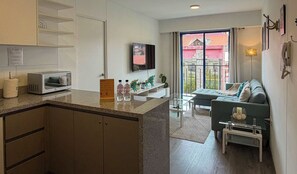 A modern kitchen with a microwave, a coffee maker, and a small refrigerator. There is a dining area with a sofa and a glass coffee table. A TV is mounted on the wall, and a balcony with a view of buildings is visible through the window. - w* | Enchanting 2BR in Historic Cusco w/ Balcony (Cusco)