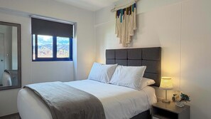 Ein Schlafzimmer mit einem Bett, einem Nachttisch, einer Lampe, einem Fenster mit Aussicht und einer Wanddekoration aus Wandteppich.