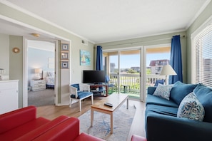 Ein Wohnzimmer mit einer Couch, einem roten Sessel, einem Fernseher und einem Balkon mit Vorhängen.