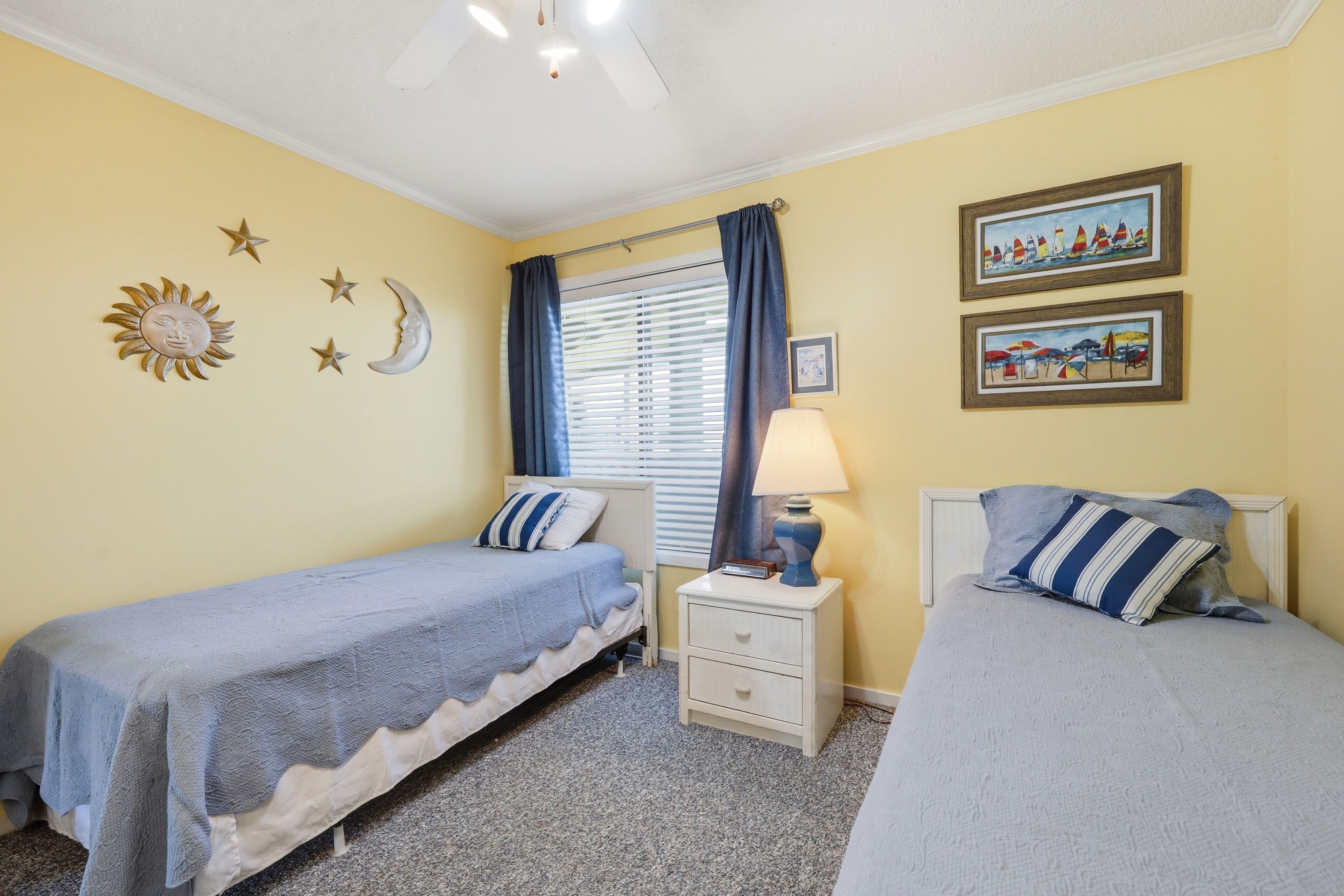 Quarto com duas camas de solteiro, mesa de cabeceira com abajur, janela com cortinas e decorações nas paredes, incluindo estrelas e um sol.