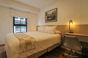 A hotel room with a bed, a desk, a chair, a window, and a wall-mounted lamp. - w* | Design-Forward 2BR w/ Balcony in Miraflores (Miraflores)