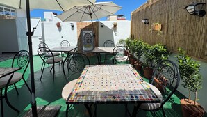 Un patio exterior con mesas y sillas, un paraguas grande, plantas en macetas y un muro decorativo.