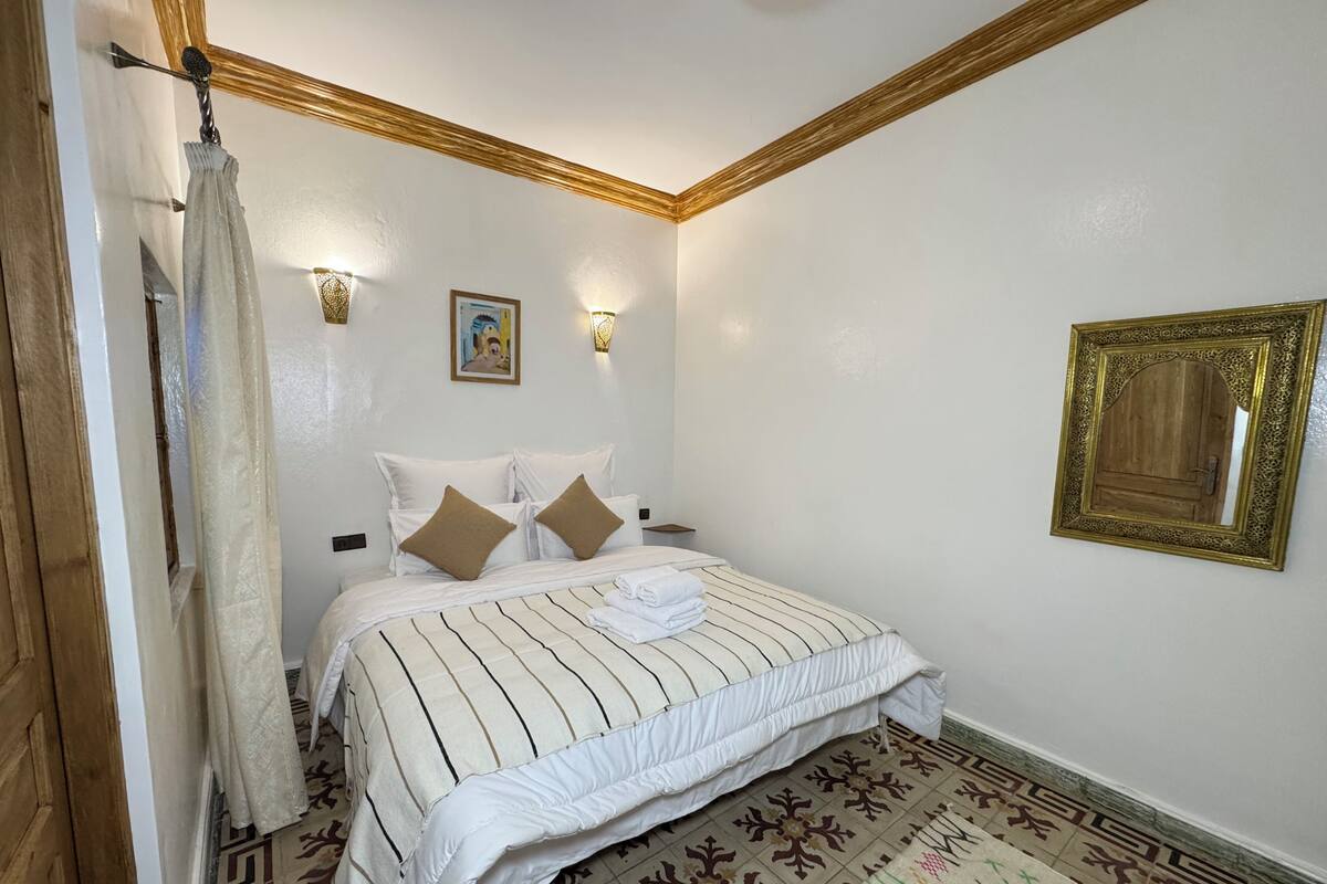 A bedroom with a bed, two wall-mounted lights, a framed mirror, and patterned floor tiles.