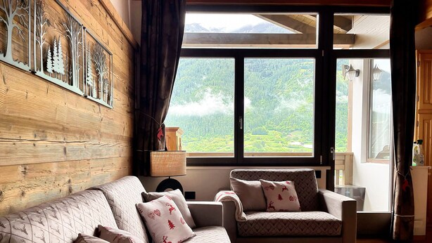 Ein gemütliches Zimmer mit Holzwand, einer Couch, einem Sessel, einem Teppich und einem großen Fenster mit Blick auf einen Wald.