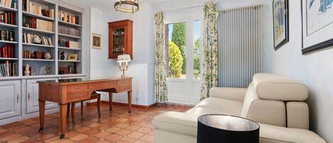 A room with a beige sofa, a wooden desk, a bookshelf, and a window with curtains.