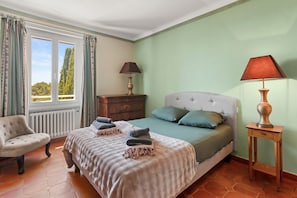 Ein Schlafzimmer mit einem Bett, zwei Nachttischen, einem Sessel, einem Fenster mit Vorhängen und Blick auf Bäume.