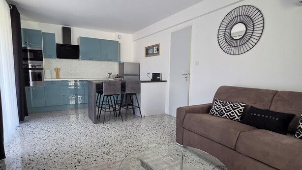 Een moderne woonkamer met een glazen salontafel, een bruine bank met gekleurde kussens, en een keuken op de achtergrond.