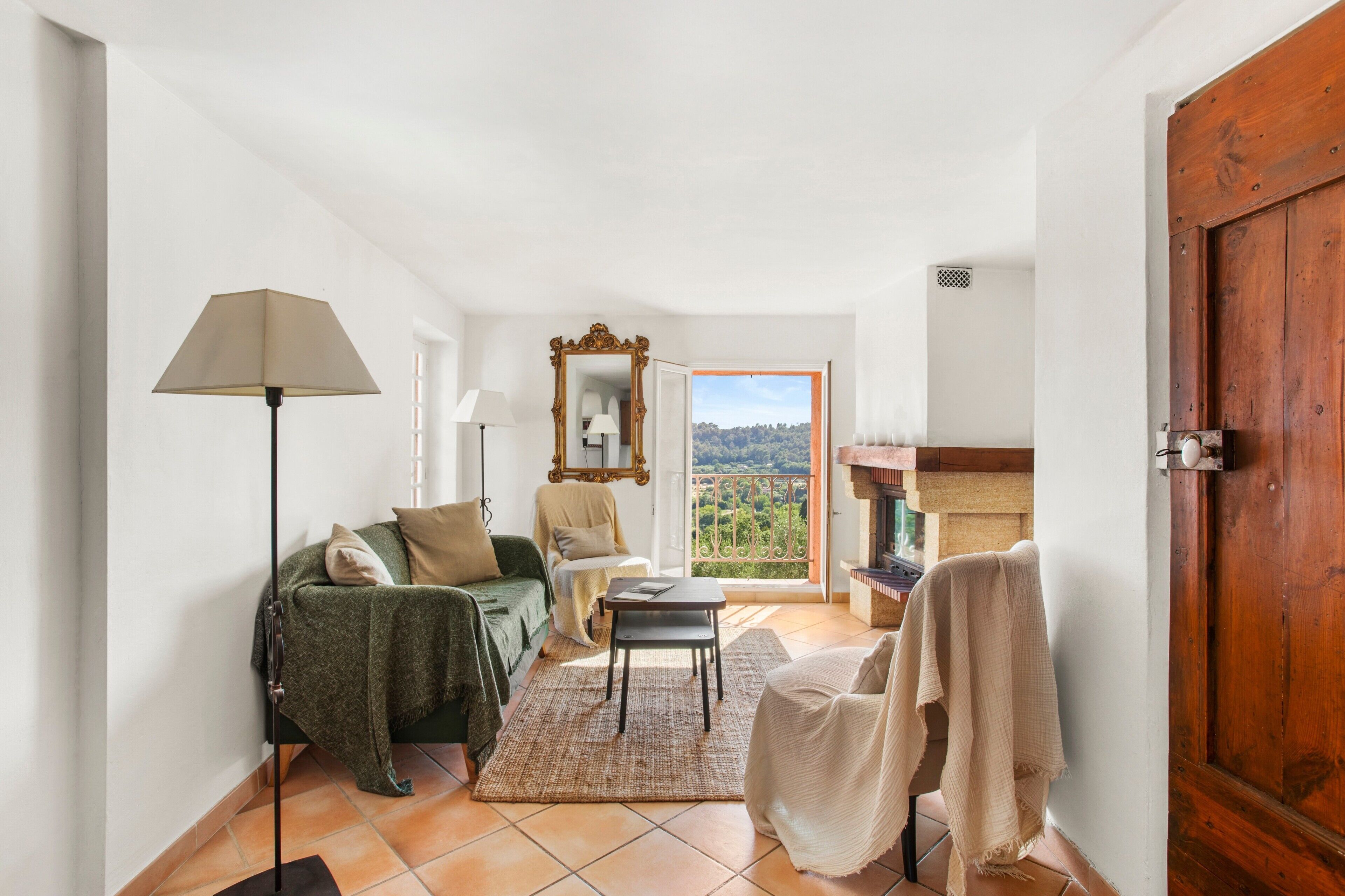 Una habitación con sofá, una silla, una mesita, una lámpara, un espejo y vistas al exterior.