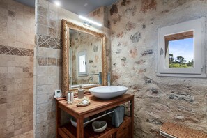 Een badkamer met een stenen muur, een grote spiegel, een houten wastafelmeubel, een witte wasbak en een raam met uitzicht.