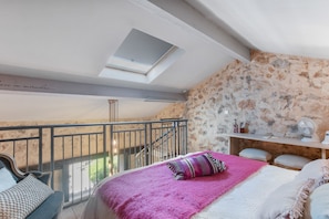 Un dormitorio con una cama, una manta rosa, un tragaluz, una escalera y una pared de piedra.