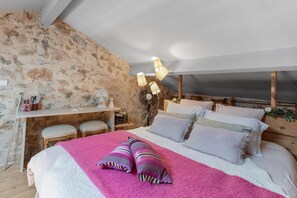 Een slaapkamer met een stenen muur, een bureau, een bed met kussens en een lamp.