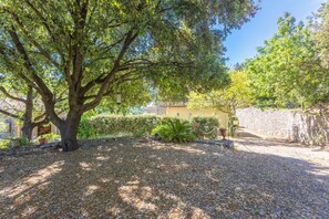 Un patio arbolado con un muro de piedra, suelo de grava y un edificio amarillo al fondo.
