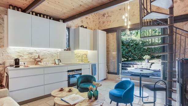 Une cuisine moderne avec des meubles blancs, un coin repas avec une table ronde et des chaises bleues, et un escalier menant à un espace extérieur.