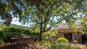 Un espace extérieur aménagé pour les repas, avec une table et des chaises, un grand arbre et un bâtiment doté d’un toit en tuiles.