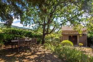 Un espace extérieur aménagé pour les repas, avec une table et des chaises, un grand arbre et un bâtiment doté d’un toit en tuiles.