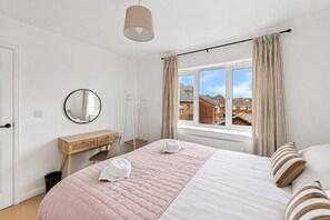 A bedroom with a bed, a desk, a chair, a window with curtains, and a view of houses outside. - The Wiltshire •  Detached 5BR House • Driveway Parking • Work Space • Smart TVs • By Yoko Property (Milton Keynes)