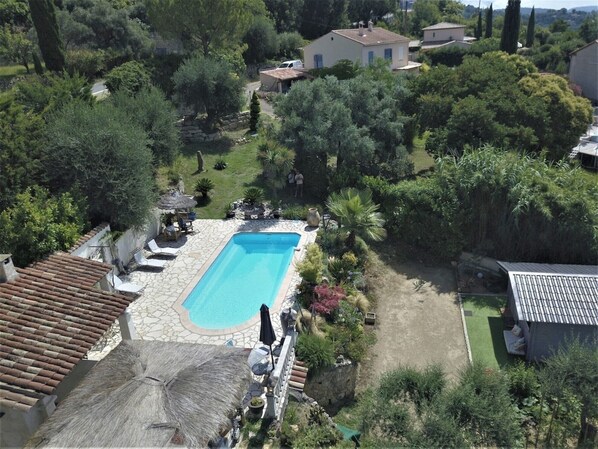 Ein Pool, umgeben von einem Garten mit einer Steinterrasse, einem strohgedeckten Gebäude und einem kleinen Bau im Hintergrund.