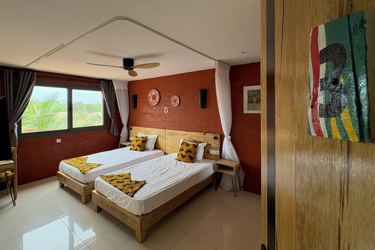 A bedroom with two beds, a ceiling fan, a window with curtains, a bedside table, and a wall-mounted artwork.