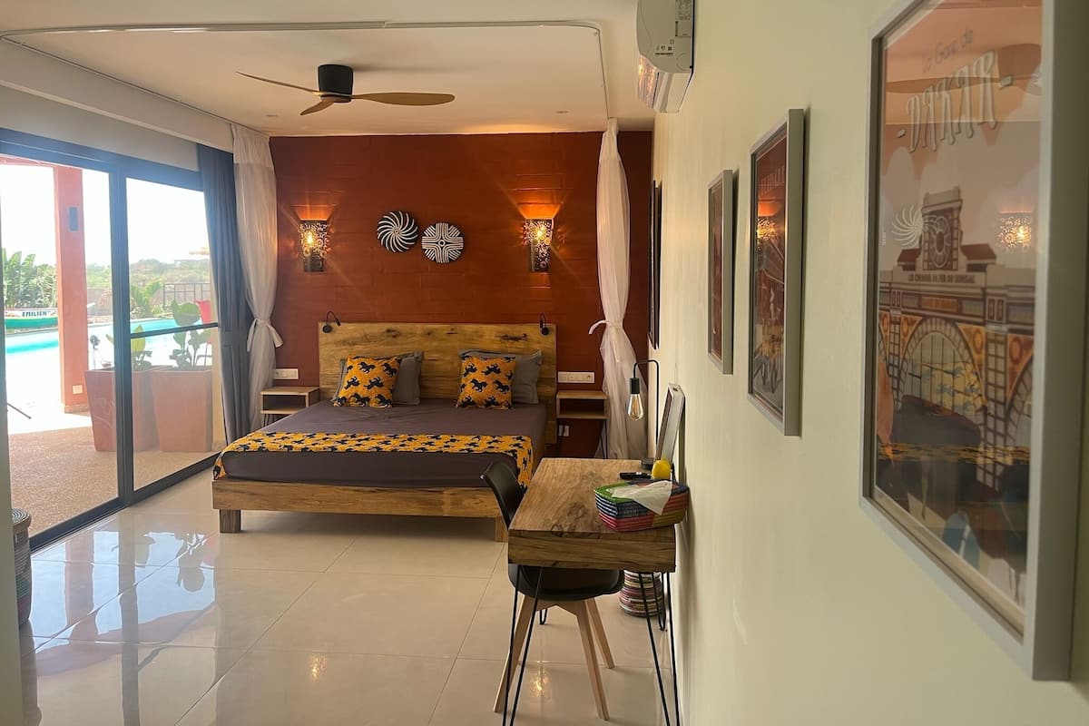 A bedroom with a bed, a desk, a ceiling fan, wall-mounted lights, and a large window.
