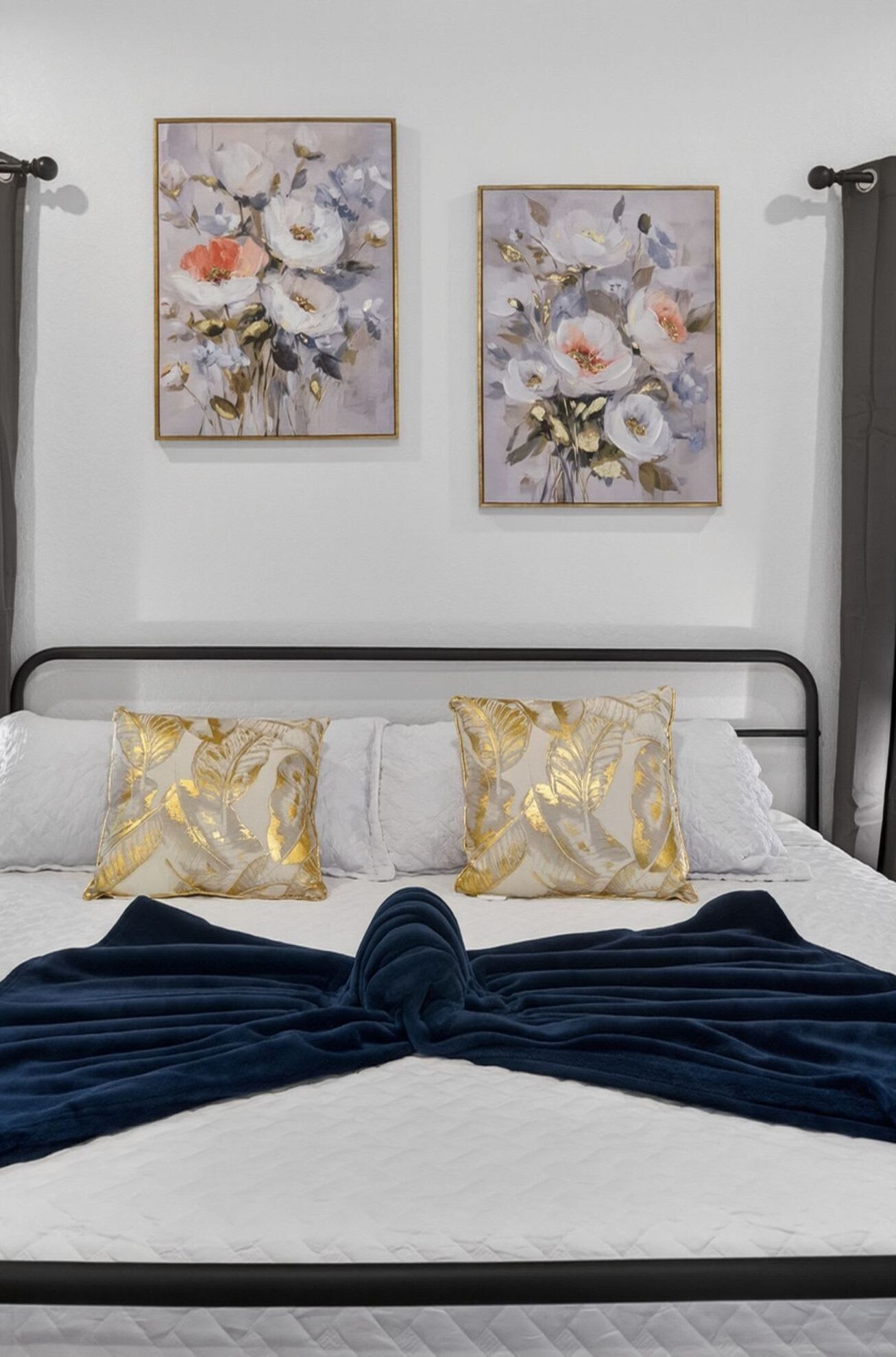 A bed with a dark blue blanket, white pillows with gold leaf patterns, and two floral paintings on the wall.