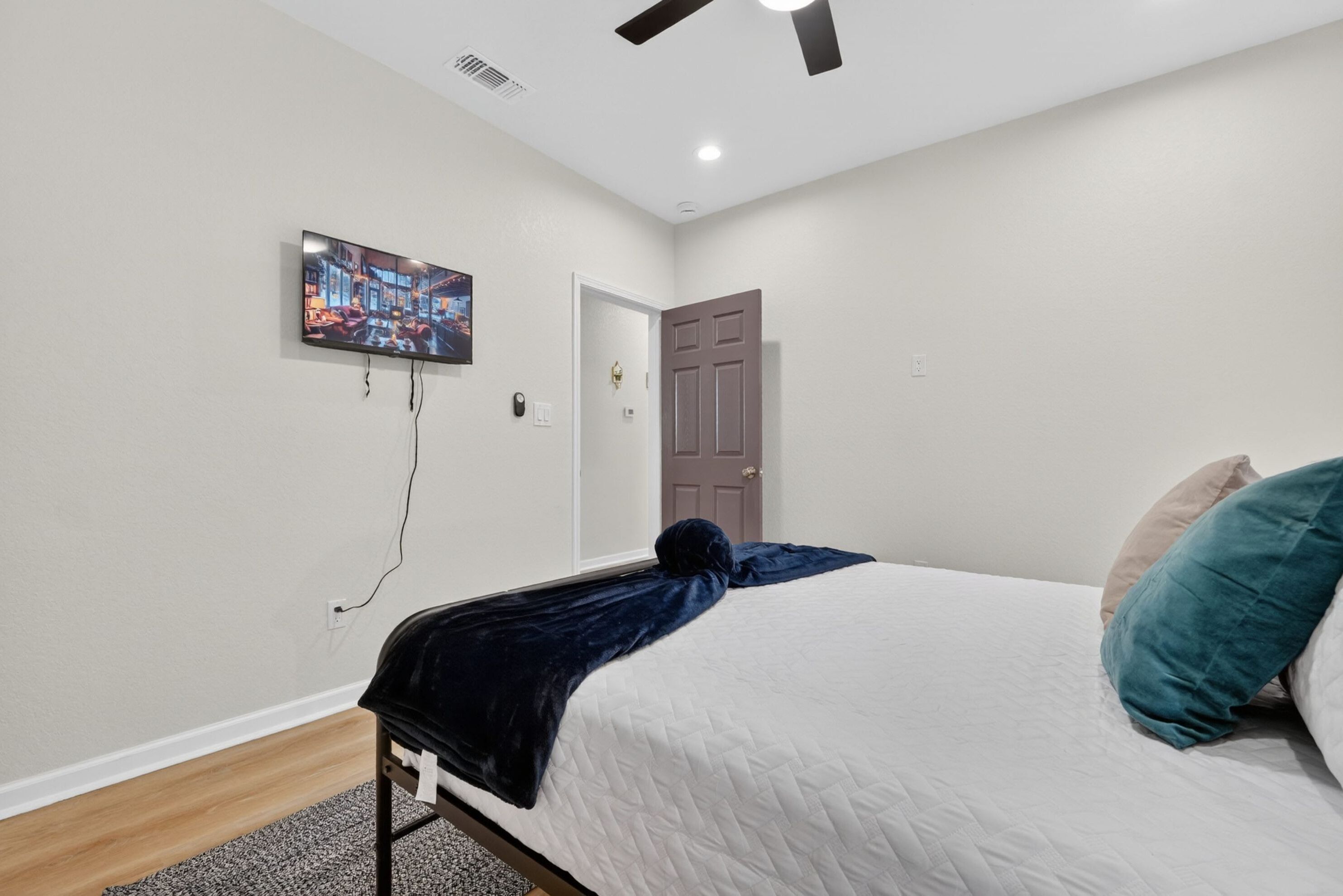 A bedroom with a bed, a TV mounted on the wall, a ceiling fan, and a door.