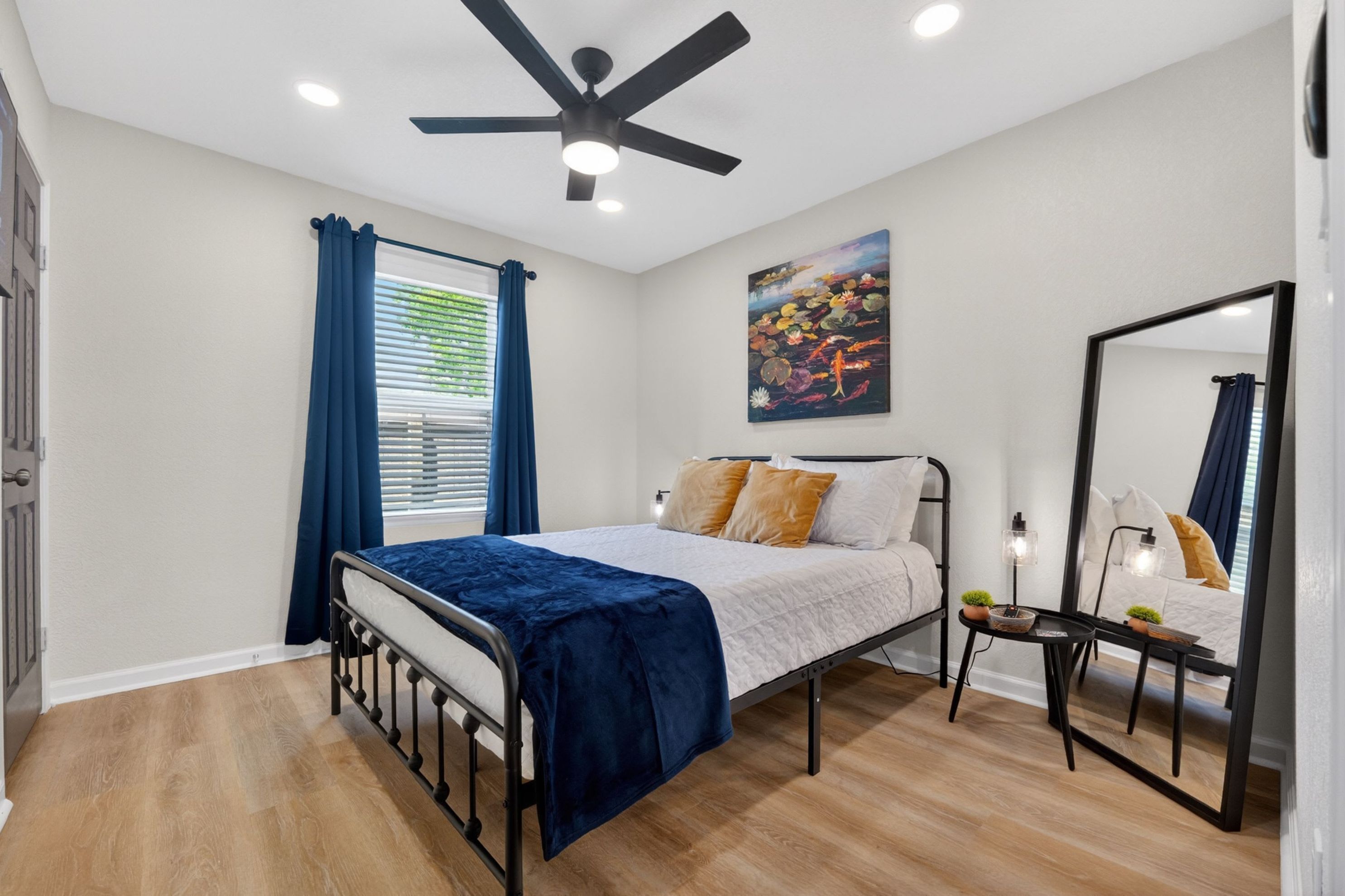 A bedroom with a bed, a ceiling fan, a window with curtains, a painting on the wall, and a mirror.