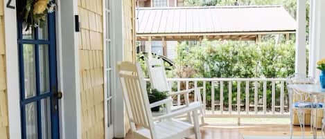 Une véranda avec une chaise berçante blanche, une décoration représentant un cygne blanc et une porte bleue portant le numéro 932.