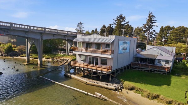 A waterfront building with a deck, a covered area, and a bridge in the background. - Two Separate Units at this Incredible Riverfront Oasis with Wraparound Deck (Florence)
