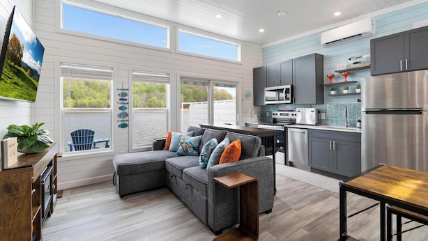 A modern kitchen with grey cabinets, a stainless steel refrigerator, and a dining area with a wooden table. - Floatopia - A Unique Floating Retreat (Russell Springs)