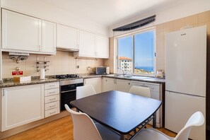 A compact kitchen with white cabinets, a stove, a sink, and a window with a view. - GueatReady - Ocean view retreat in Caniço (Caniço)