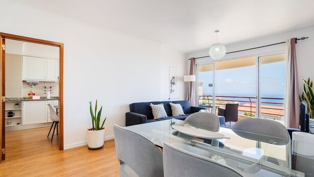 Una sala de estar moderna con una mesa de comedor de vidrio, sillas grises, un sofá azul oscuro con cojines estampados y una planta en maceta.
