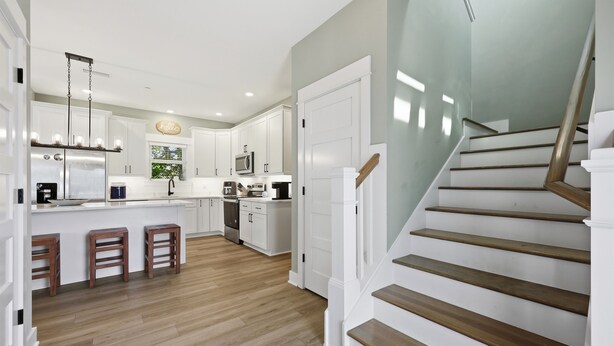 A modern kitchen with white cabinets, a stainless steel refrigerator, wooden stools, and a staircase with wooden steps. - Steps to Beach! Hot Tub, Dogs OK, Pool, Pickleball (Corolla)