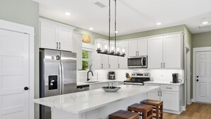 A modern kitchen with white cabinets, stainless steel appliances, a marble island, and wooden stools. - Steps to Beach! Hot Tub, Dogs OK, Pool, Pickleball (Corolla)