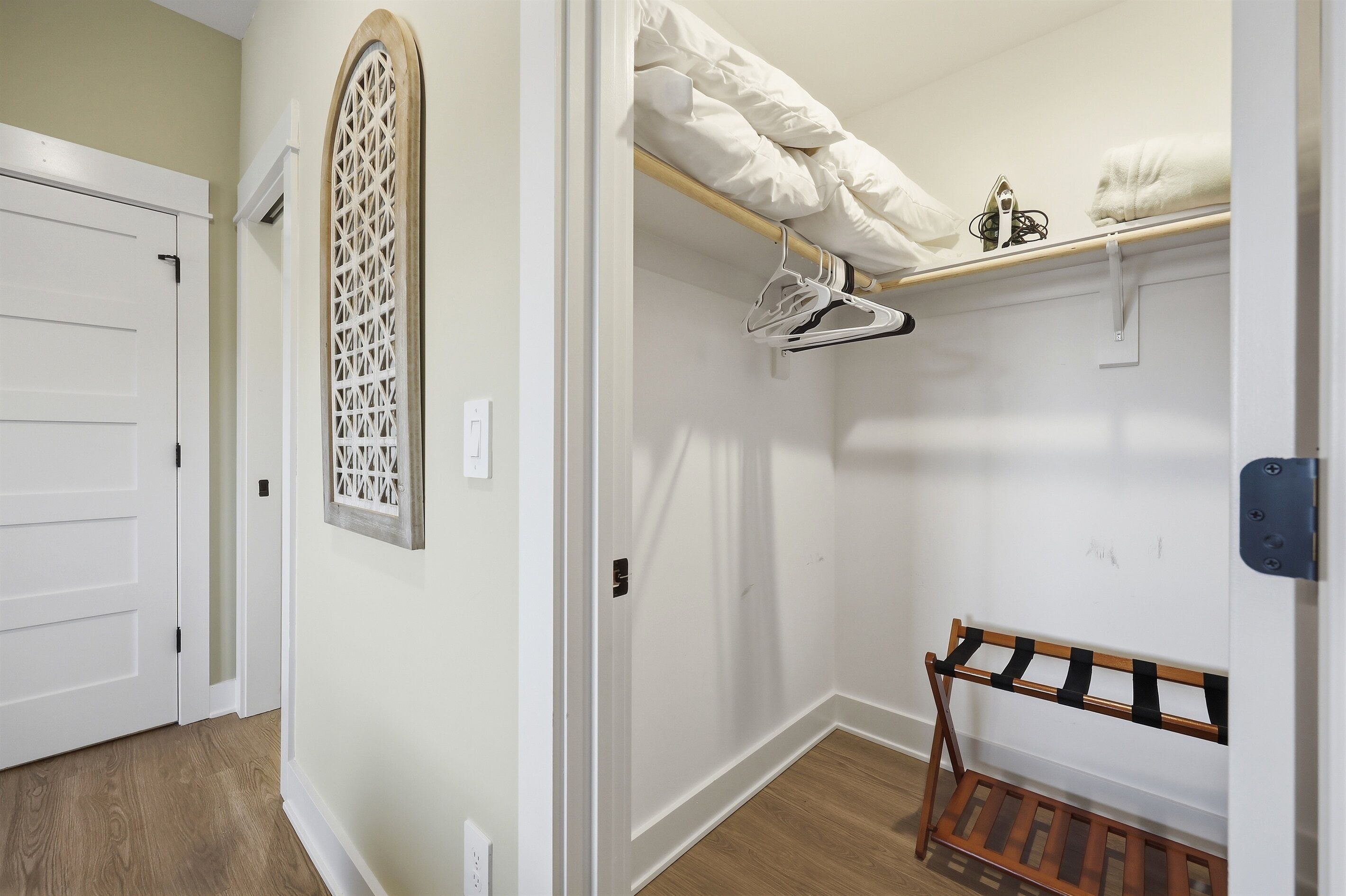 A hallway with a wooden floor, a white door, a decorative window, and a closet with a wooden rack and hanging clothes.