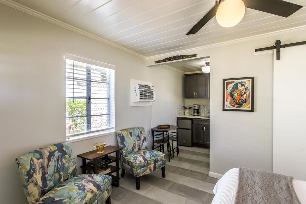 A room with a sofa, two chairs, a small table, a ceiling fan, a window, a kitchen area, and a framed picture on the wall.