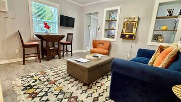 Una sala de estar con un sofá azul, un sillón naranja, un reposapiés marrón, un tapete estampado y un televisor en la pared.