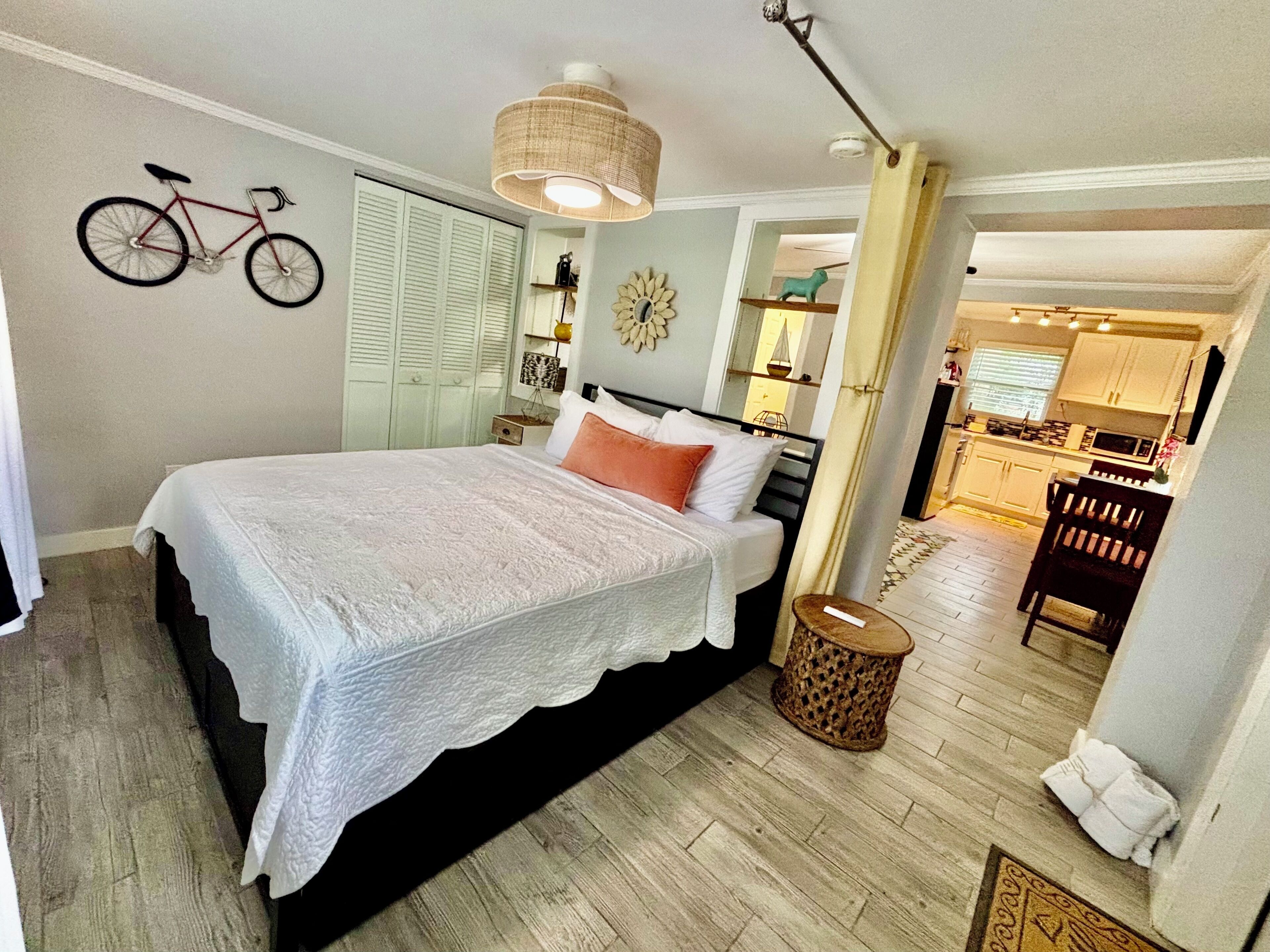 A bedroom with a bed, a bicycle on the wall, a small round table, and a view into the kitchen.
