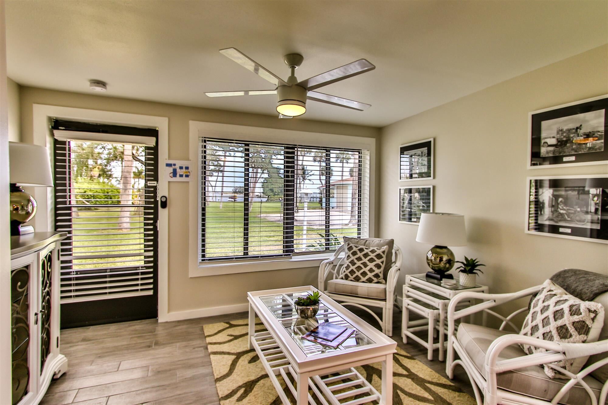 A room with a ceiling fan, a glass-top coffee table, white wicker furniture, and framed pictures on the wall.