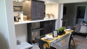 Una cocina compacta con armarios oscuros, una mesa de comedor de vidrio y una pequeña cama para mascotas.