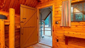Interior de uma cabana de madeira com uma porta grande, uma janela com cortinas e um banco de madeira.