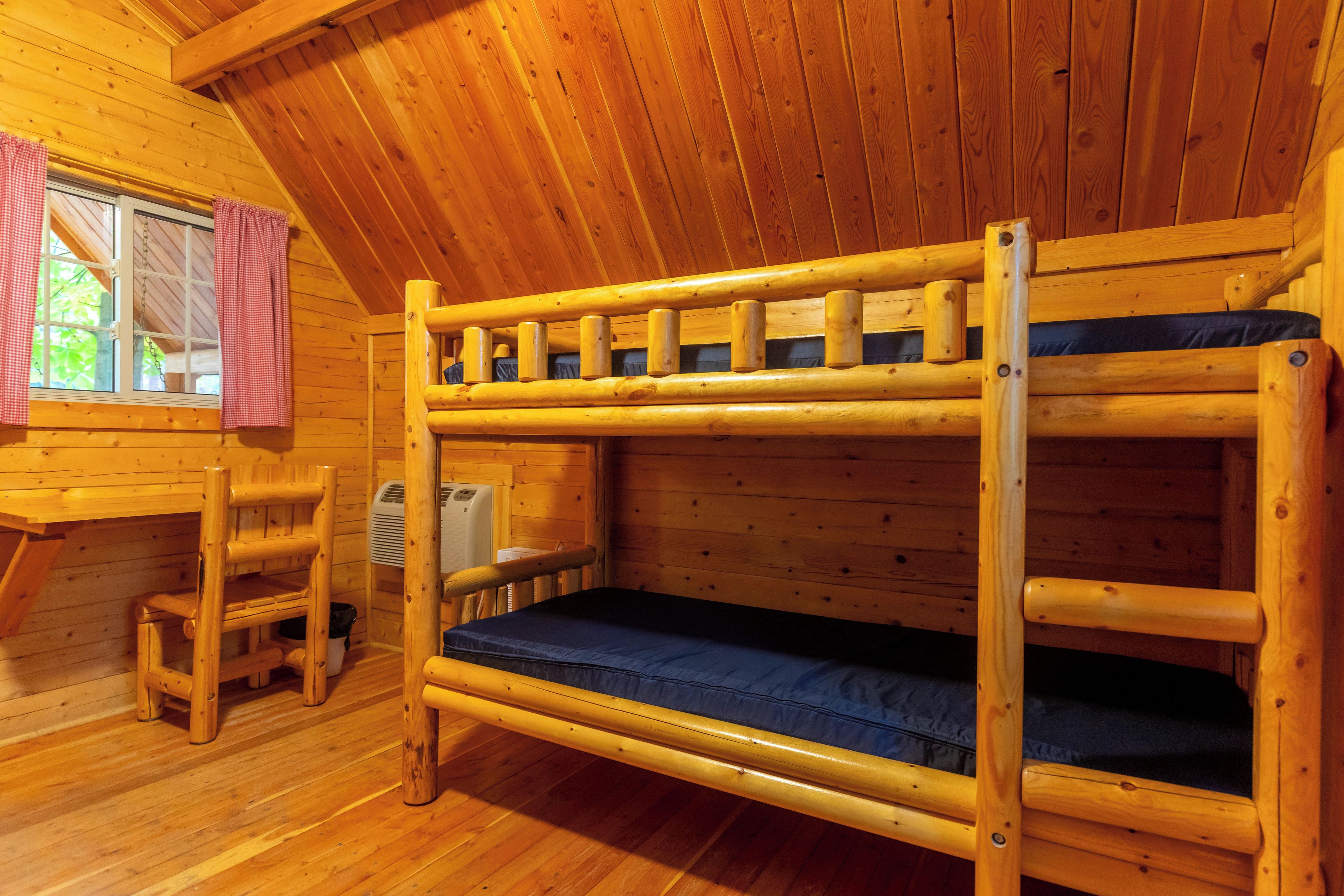 A bunk bed with a wooden frame and blue mattress, a small wooden chair, a heater, and a window with red curtains.