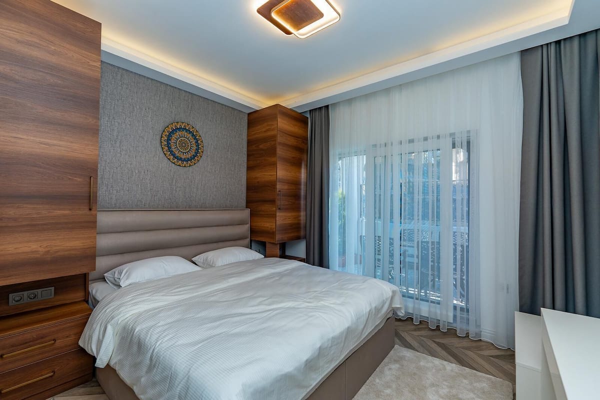 Un dormitorio moderno con una cama grande, cabecera de madera, un adorno en la pared y vistas a través de cortinas translúcidas.